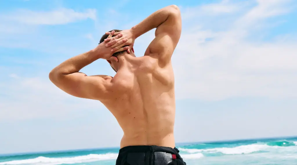 Um homem musculoso visto de costas enquanto se alonga com as mãos atrás da cabeça. Ele está em uma praia, com ondas e um céu azul ao fundo. A imagem destaca suas costas fortes e bem definidas.